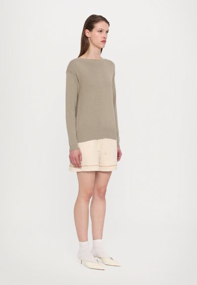 Καφέ πλεκτό πουλόβερ με ριχτή ύφανση, συνδυασμένο με μπεζ σορτς που έχει οριζόντια ρίγα, και λευκές κάλτσες με υπόδηση σε στυλ μυτερού παπουτσιού.