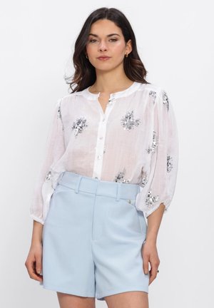 Vrouw draagt een doorschijnende witte blouse met zilveren bloemenpailletten en lichtblauwe, hoog getailleerde, op maat gemaakte shorts, staand tegen een effen achtergrond.