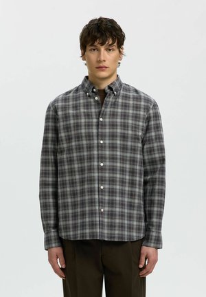 Jeune homme aux cheveux courts, bouclés et foncés, portant une chemise grise à carreaux à manches longues boutonnée et un pantalon sombre, debout devant un fond uni.
