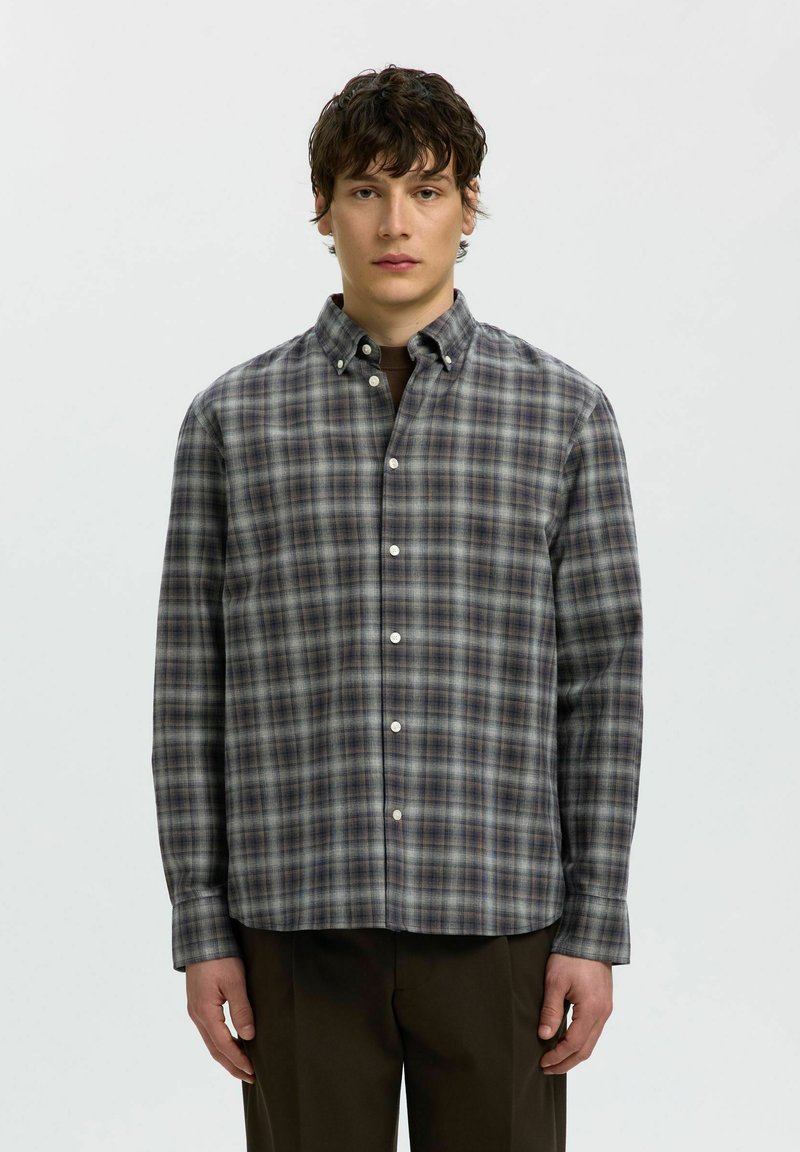 Jeune homme aux cheveux courts, bouclés et foncés, portant une chemise grise à carreaux à manches longues boutonnée et un pantalon sombre, debout devant un fond uni.