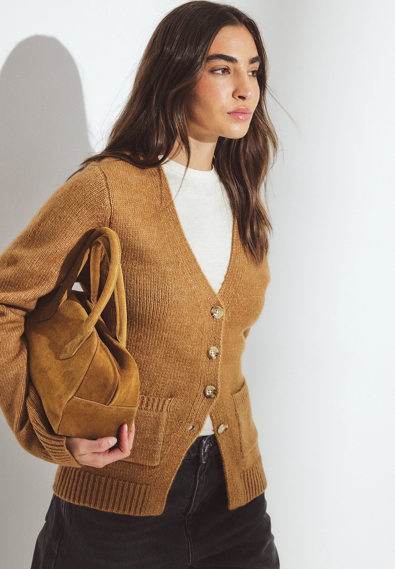 Brun strikket cardigan med V-hals, to forlommer og gyldne knapper; kombineret med en blød cremefarvet skjorte og en brun ruskindtaske.
