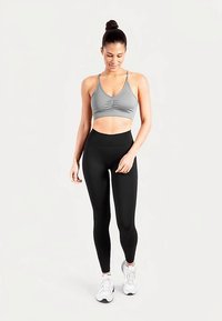 Brassière de sport grise avec un centre froncé et des bretelles fines, associée à un legging noir taille haute et des chaussures de sport blanches.