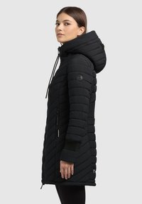 Kobieta w czarnej, pikowanej, długiej zimowej kurtce z kapturem, z zasuwanymi kieszeniami i prążkowanymi mankietami, stojąca na boku na szarym tle.