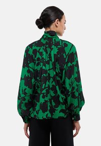 Blouse florale verte avec des motifs noirs, dotée d'un col montant, de manches ballons et d'une coupe décontractée, fabriquée en tissu léger.