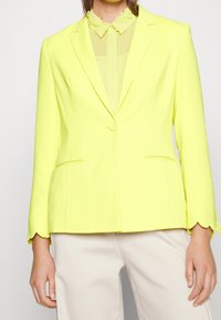 Blazer jaune avec une coupe ajustée, présentant un ourlet festonné aux manches, deux poches avant et une fermeture par bouton. En dessous se trouve un chemisier transparent.
