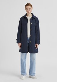 Marineblauer, wasserabweisender Mantel mit Kragen, Knopfverschluss und zwei seitlichen Taschen. Kombiniert mit hellen Jeans und weißen Turnschuhen.