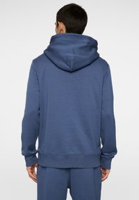 Blå sweatshirt med stor huva, ribbade ärmmuddar och nederkant. Slät textur med minimala designdetaljer; visad bakifrån.