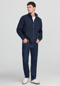Giacca blu navy con zip e colletto alto. Realizzata in materiale morbido, presenta tasche laterali ed è abbinata a jeans scuri e sneakers bianche.