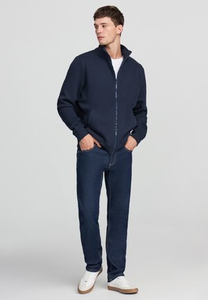 Giacca blu navy con zip e colletto alto. Realizzata in materiale morbido, presenta tasche laterali ed è abbinata a jeans scuri e sneakers bianche.