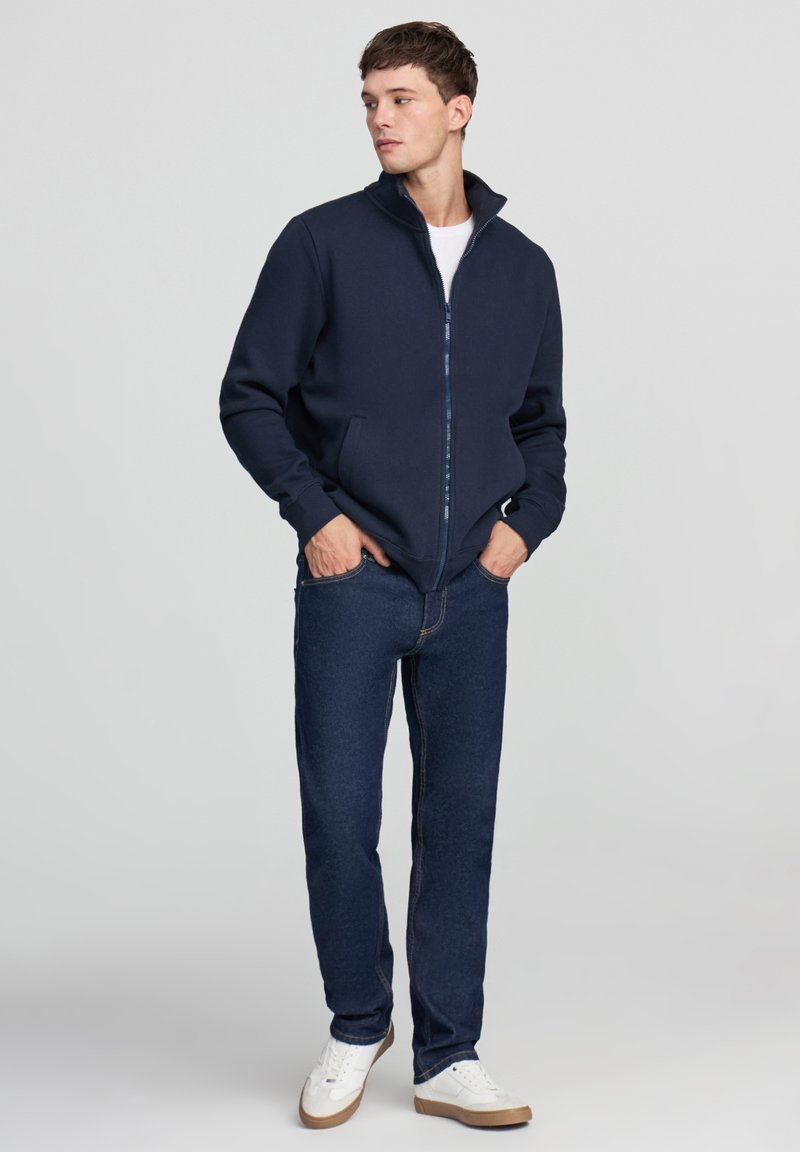 Giacca blu navy con zip e colletto alto. Realizzata in materiale morbido, presenta tasche laterali ed è abbinata a jeans scuri e sneakers bianche.