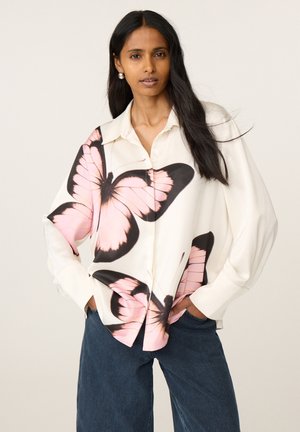 Vrouw met lang donker haar draagt een wit overhemd met grote roze en zwarte vlinderprints en donkerblauwe wijdvallende spijkerbroeken.