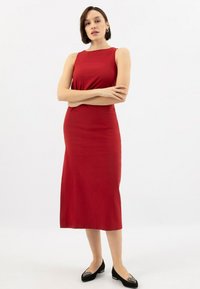 Donna con capelli corti scuri che indossa un vestito midi rosso senza maniche e scarpe basse nere con decorazioni, in piedi con le braccia incrociate.