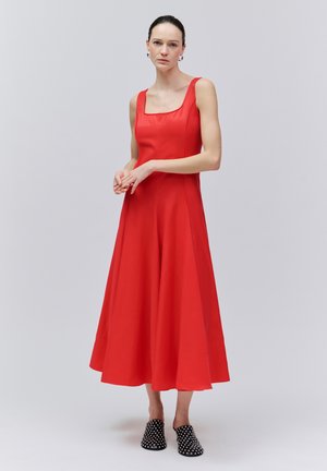 Žena v červenom šatách bez rukávov so štvorcovým výstrihom a čiernych šľapkách s cvočkami, stojaca pred jednoduchým svetlosivým pozadím.