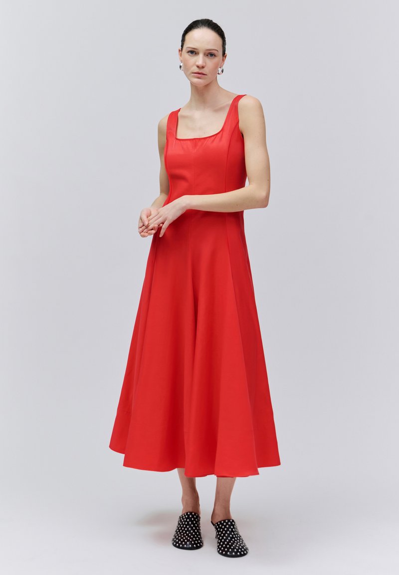 Femme portant une robe midi rouge sans manches avec un décolleté carré et des chaussures slip-on noires cloutées, debout devant un fond gris clair uni.
