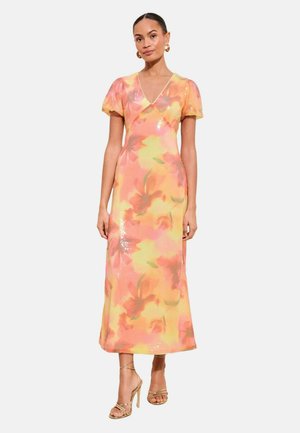 Kobieta ubrana w sukienkę midi z cekinami w pastelowym różu i żółci z kwiatowym wzorem, z bufkami na rękawach, na nogach złote, cienkie szpilki, stojąca na białym tle.