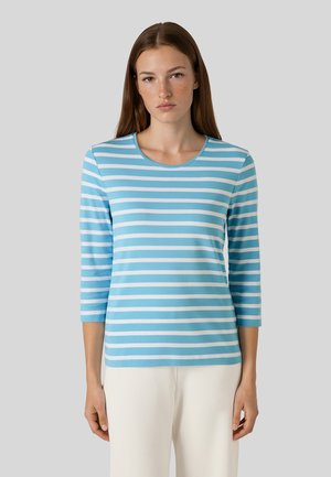 Vrouw met lang bruin haar die een lichtblauw-wit horizontaal gestreept shirt met driekwart mouwen en witte broek draagt.