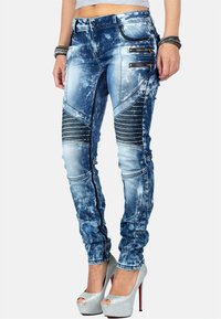 Pantaloni in denim blu aderenti con finitura usurata, accenti texture, pannelli a coste e dettagli con zip, abbinati a tacchi alti argentati.