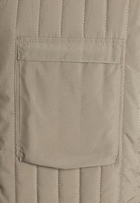 Beige gesteppter Stoff mit einer aufgesetzten Tasche. Die Textur ist glatt mit einem dezenten Glanz, und die Nähte sorgen für eine strukturierte Optik.