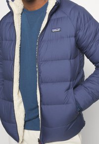 Marinblå pufferjacka med en krämfärgad faux fur-foder, dragkedjestängning och sidofickor. Ytmaterialet verkar slätt och något glänsande.