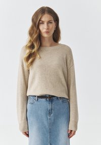 Beige gebreide trui met een geribbelde ronde halslijn en lange mouwen, gecombineerd met een lichtblauwe spijkerrok. De trui heeft een casual pasvorm.