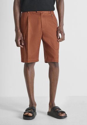 Man die roestkleurige cargoshorts en zwarte open teen sandalen draagt, staand op een effen lichte achtergrond.