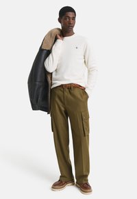Weißer Pullover, olivfarbene Cargo-Hosen mit Taschen, brauner geflochtener Gürtel und braune Lederschuhe. Schwarze Lederjacke mit Fellfutter.