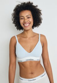 Svetlo modra bralette s globokim V-izrezom, nastavljivimi naramnicami in belim pasom, na katerem je v črnih črkah napisano "Calvin Klein" ob dnu.