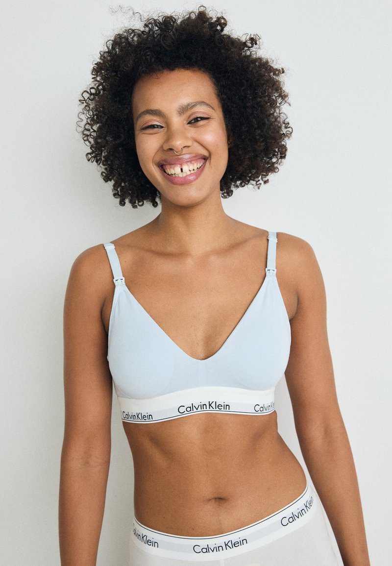 Svetlo modra bralette s globokim V-izrezom, nastavljivimi naramnicami in belim pasom, na katerem je v črnih črkah napisano "Calvin Klein" ob dnu.