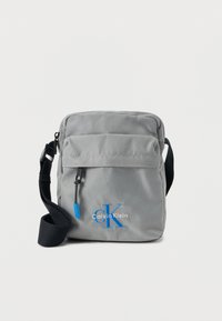 Γκρι τσάντα ώμου από νάιλον με φερμουάρ, ρυθμιζόμενο λουρί και μπλε λεπτομέρειες λογότυπου. Διαθέτει μπροστινή τσέπη με μεταλλικά στοιχεία.