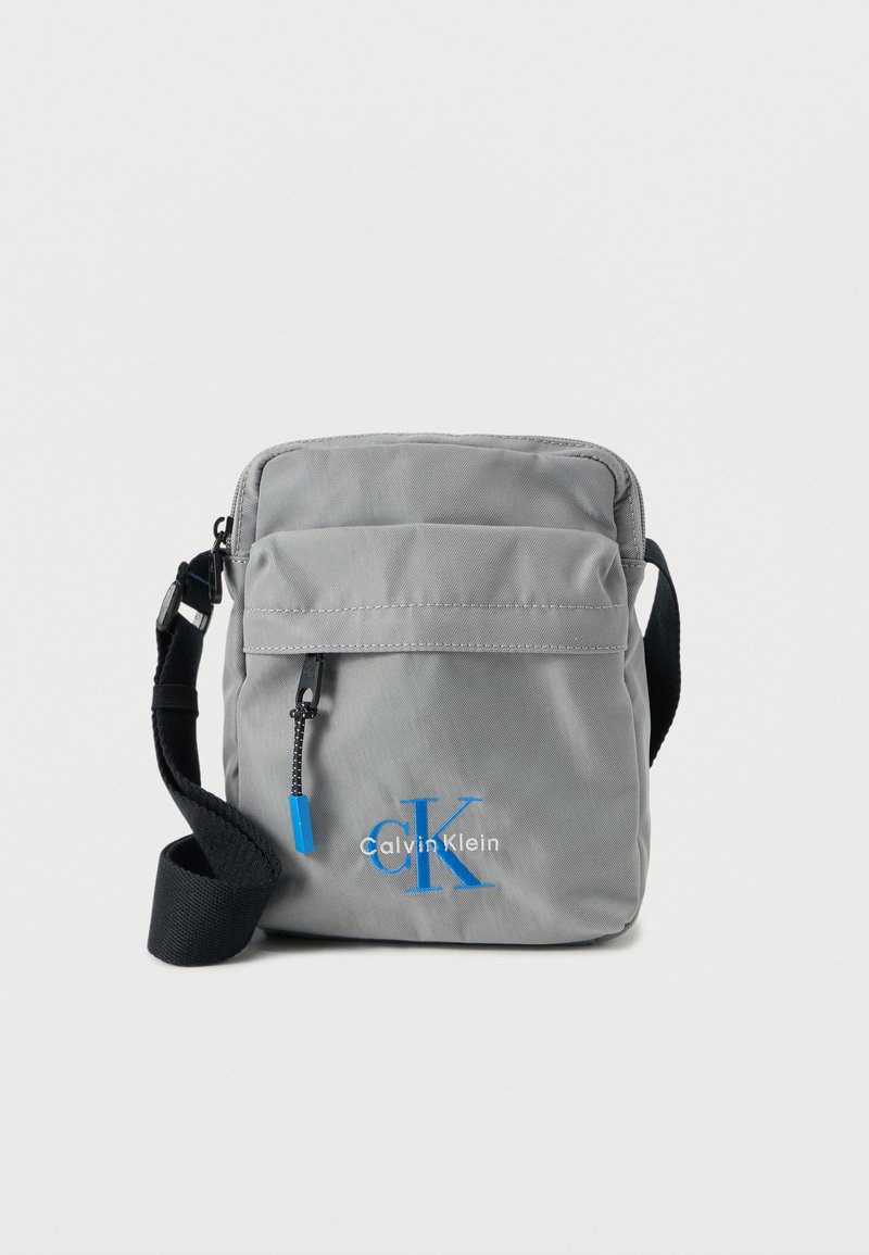 Γκρι τσάντα ώμου από νάιλον με φερμουάρ, ρυθμιζόμενο λουρί και μπλε λεπτομέρειες λογότυπου. Διαθέτει μπροστινή τσέπη με μεταλλικά στοιχεία.