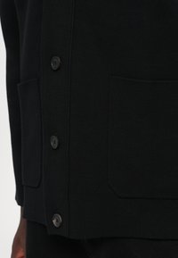 Crni kardigan od rebraste pletenine, s četiri gumba, dva prednja džepa i ravnim rubom. Uočena teksturirana površina.