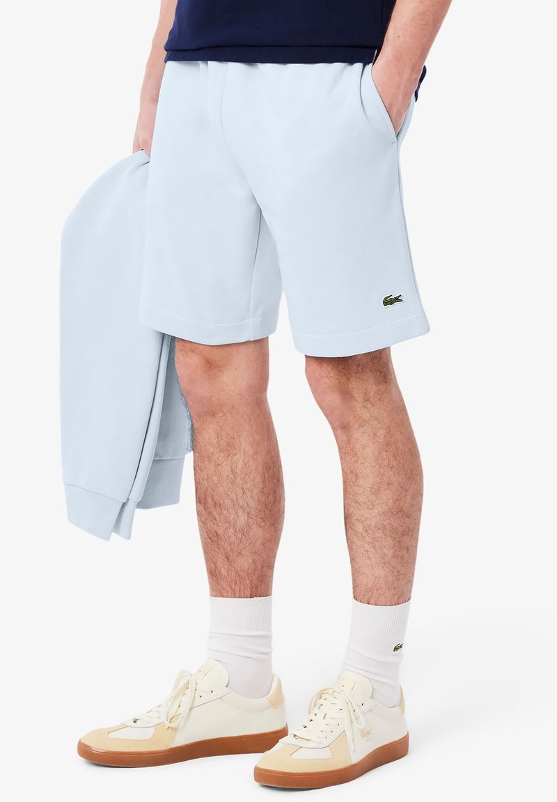 Shorts en coton bleu clair avec une coupe décontractée et un petit logo vert sur l'ourlet. Associés à des baskets crème et des chaussettes blanches.