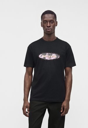 Homme debout face à l'avant portant un t-shirt noir avec un logo graphique rose et blanc au centre.