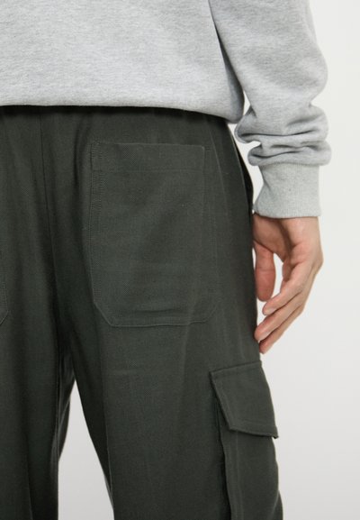 Cargo pants in dark green fabric featuring two large front pockets and visible stitching. The waistband appears elasticized for comfort.