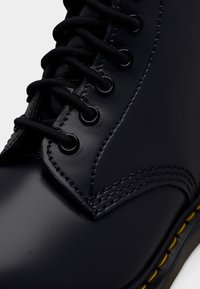 Sort læderstøvle med rund tå, tekstureret overflade, sorte huller og snørebånd. Har gul syning langs sålen for kontrast.