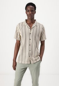 Chemise à manches courtes avec boutons, beige à rayures noires et accents à pois. Tissu doux avec une coupe décontractée. Portée avec un pantalon vert clair.