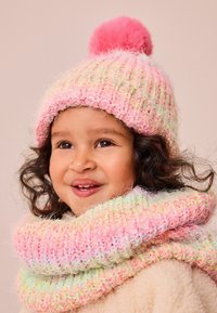 Gehaakte roze muts met een fluffige roze pompon en kleurengaren; bijpassende chunky sjaal om de nek gewikkeld; beide hebben een zachte textuur.