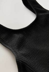 Top in maglia nera con scollatura ampia e tessuto testurizzato. Le spalline sono spesse e il design complessivo è semplice ma moderno.