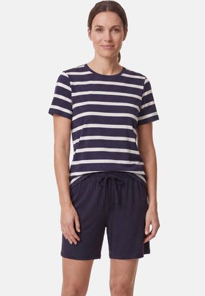 Vrouw draagt een marineblauw-wit gestreept T-shirt met korte mouwen en marineblauwe short met trekkoord, staand tegen een effen lichte achtergrond.