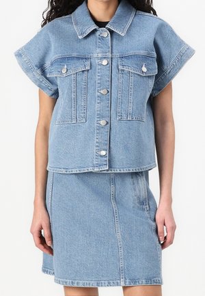 Vrouw draagt een lichtblauw denim jack met korte mouwen en zilverkleurige knopen, gecombineerd met een bijpassende denim rok tot op de knieën.