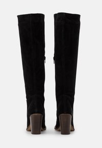 Botas de ante negras hasta la rodilla con puntera redonda, detalles de costura vertical y un tacón de madera grueso. Acento de cremallera en el interior.