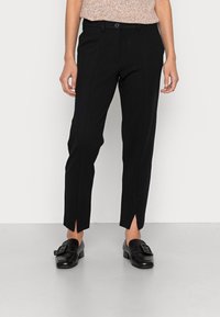 Pantalon noir slim fit en tissu lisse, avec coutures verticales et fentes latérales à l'ourlet. Associé à des mocassins noirs.