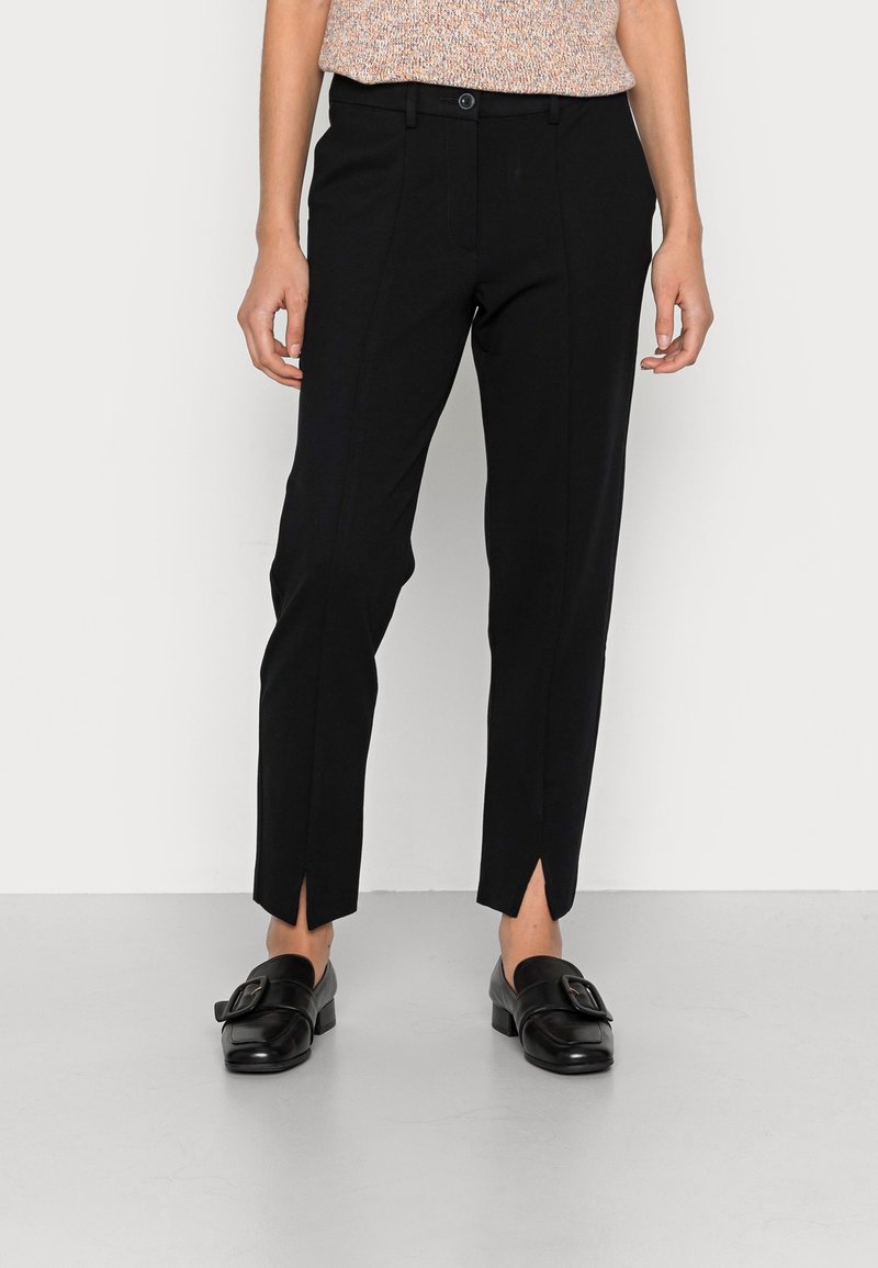 Pantalon noir slim fit en tissu lisse, avec coutures verticales et fentes latérales à l'ourlet. Associé à des mocassins noirs.