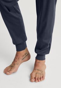 Donkerblauwe joggingbroek met elastische boorden, met een gladde textuur. Blootsvoets met verschillende zwarte tatoeages zichtbaar.
