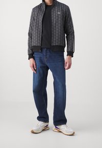 Homme portant une veste noire zippée à motifs, une chemise noire, un jean bleu et des baskets blanches avec des accents violets et beige, debout sur un fond blanc.