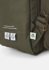 Green recycled fabric backpack featuring a metal buckle, labels for materials used, and a textured surface. "This bag was trash!" printed prominently.