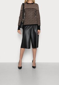Schwarze Kunstleder-Culottes mit lockerem Schnitt, kombiniert mit einem gestreiften Strickpullover, der das Wort "GUESS" in wechselnden Farben und mit goldenen Knopf-Details zeigt.
