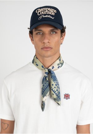 Mann trägt eine marineblaue Kappe mit dem Text "Champagne Courtside Prohibited", ein weißes T-Shirt mit rotem Logo und einen gemusterten blau-beigen Halstuch, vorne gebunden.