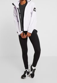 Persona vistiendo una chaqueta blanca de Adidas, leggings negros, zapatillas en blanco y negro, de pie contra un fondo blanco liso.