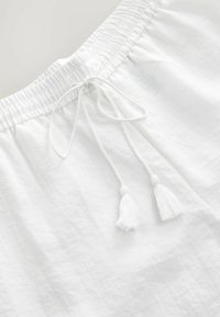 Shorts blancs en lin avec une taille élastique, cordon de serrage ajustable avec deux pompons blancs, et une surface texturée.